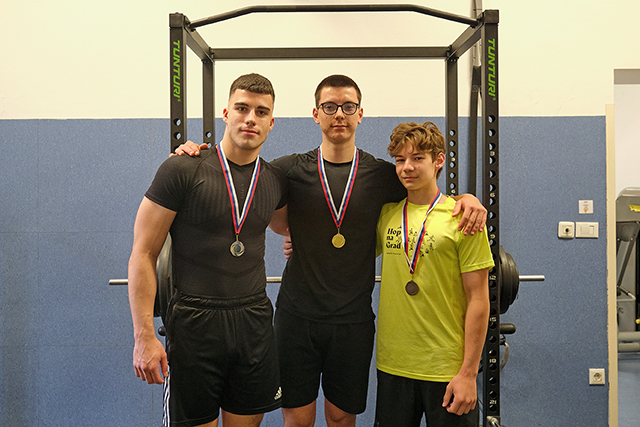 Na sliki so trije fantje, ki so tekmovali v dvigovanju uteži. Objeti so čez ramena, okrog vratu nosijo medalje za dosežena mesta.