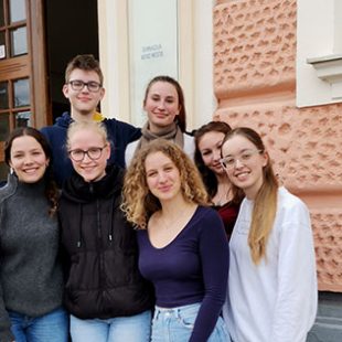 Šest dijakinj in dijak stojijo na stopnišču pred Gimnazijo Novo mesto, kjer je potekalo državno tekmovanje iz znanja psihologije. Zrejo v kamero, so nasmejani.