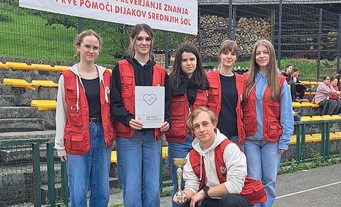 Slika prikazuje pet dijakinj in dijaka. Dijakinje stojijo, dijak pa čepi. V rokah drži pokal, ena od dijakinj pa priznanje za 4. mesto na tekmovanju iz znanja prve pomoči. Vsi so oblečeni v rdeče rešvalske brezrokavnike.
