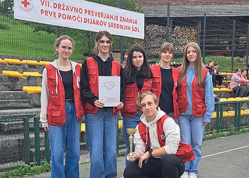 Slika prikazuje pet dijakinj in dijaka. Dijakinje stojijo, dijak pa čepi. V rokah drži pokal, ena od dijakinj pa priznanje za 4. mesto na tekmovanju iz znanja prve pomoči. Vsi so oblečeni v rdeče rešvalske brezrokavnike.