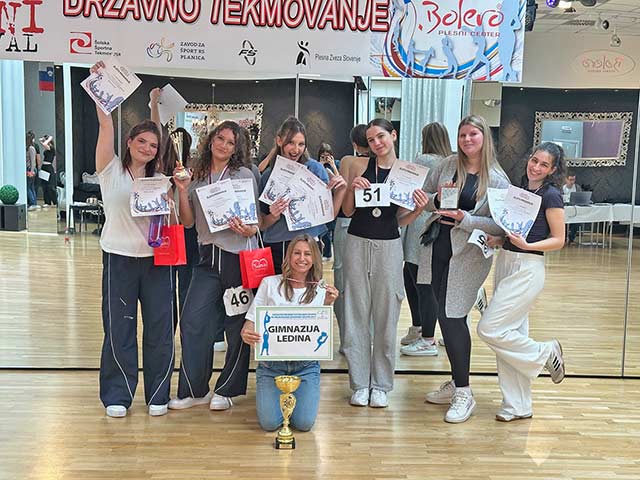Šest plesalk drži v rokah priznanja, okrog vratu imajo medalje, pred njimi na tleh sedi mentorica in z eno roko drži medaljo, z drugo napis "Gimnazija Ledina". Pred njo na tleh je pokal.