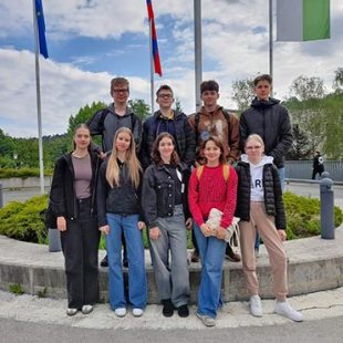 Devet dijakov, udeležencev kemijskega tekmovanja, stoji pred zelenico pred Fakulteto za kemijo in keimijsko tehnologijo. V ozadju so drogovi z zastavami.