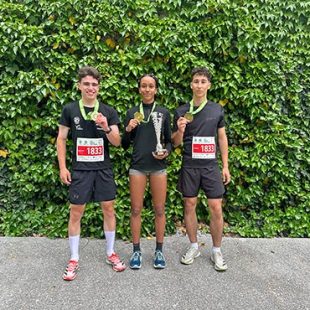Dva dijaka in dijakinja stojijo pred steno obraščeno z bršljanom. Okrog vratu imajo medalje za 1. mesto na teku trojk.