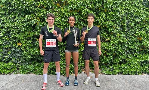 Dva dijaka in dijakinja stojijo pred steno obraščeno z bršljanom. Okrog vratu imajo medalje za 1. mesto na teku trojk.