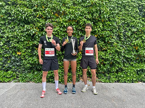Dva dijaka in dijakinja stojijo pred steno obraščeno z bršljanom. Okrog vratu imajo medalje za 1. mesto na teku trojk.