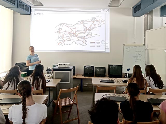 Dijaki sedijo v učilnici in poslušajo predavanje o razvoju interneta v Sloveniji. Na projekcijskem platnu na steni so prikazane glavne internetne žile v Sloveniji.
