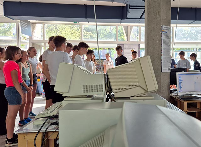 Fotografija prikazuje skupino dijakov v muzeju. Pred njima sta dva računalniška monitorja iz 90. let. Monitorji iz tistega časa niso bili tanko kot današnji, ampak so imeli obliko škatle.