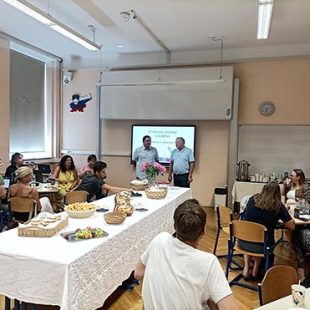 Na fotografiji je dolga pogornjena miza s prigrizki, levo in desno od nje sedijo udeleženci srečanja Katarina. Pred projekcijskim platnom stojita ravnatelj I. gimnazije v Celju in profesor Andrej Brodnik. Imata uvodni nagovor.