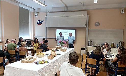 Na fotografiji je dolga pogornjena miza s prigrizki, levo in desno od nje sedijo udeleženci srečanja Katarina. Pred projekcijskim platnom stojita ravnatelj I. gimnazije v Celju in profesor Andrej Brodnik. Imata uvodni nagovor.