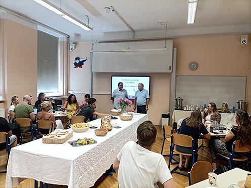 Na fotografiji je dolga pogornjena miza s prigrizki, levo in desno od nje sedijo udeleženci srečanja Katarina. Pred projekcijskim platnom stojita ravnatelj I. gimnazije v Celju in profesor Andrej Brodnik. Imata uvodni nagovor.