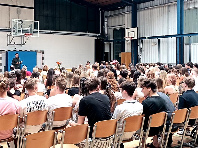 Maturantje sedijo na stolih v telovadnici, kjer poteka podelitev maturitetnih spričeval. V ozadju fotografije je ravnatelj med govorom.