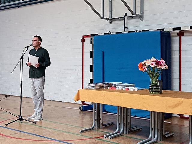 Ravnatelj stoji pred mikrofonom in ima govor na podelitvi maturitetnih spričeval. Na mizi poleg njega je vaza z rožami in nagrade za zlate ter diamantne maturante.