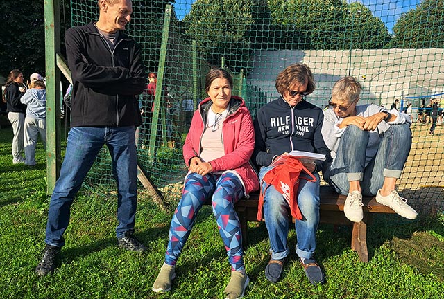 Profesorji spremljajo športne dejavnosti in seštevajo rezultate. Na klopci sedijo dve profesorici in profesor, ki si rezultate zapisuje na list, poleg njih stoji drugi profesor, ki si ogleduje zapisano.