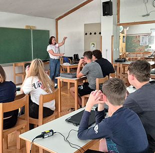 Dijaki so v učilnici, na mizah imajo prenosne računalnike. Poslušajo razlago učiteljice.