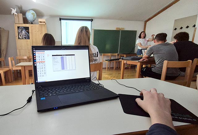 Na mizi je odprt prenosni računalnik s tabelo na zaslonu, v ozadju fotografije pa je videti druge dijake in učiteljico.