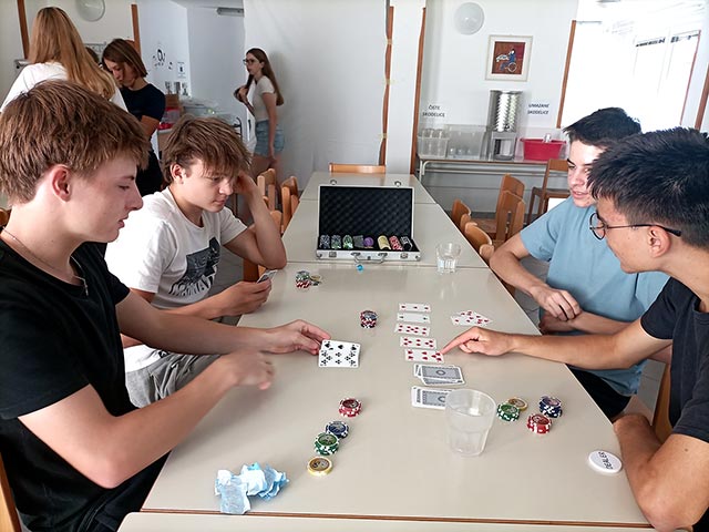 Štirje dijaki sedijo za mizo in igrajo poker. Poleg kart so na mizi tudi pisani žetoni za pobiranje stav.