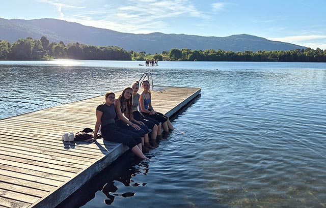 Štiri dijakinje sedijo na lesenem pomolu. Noge namakajo v jezero. Dan je sončen.