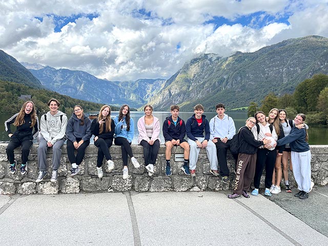 Dijaki sedijo na kamniti ograji mostu, v ozadju je Bohinjsko jezero z okoliškini hribi. 