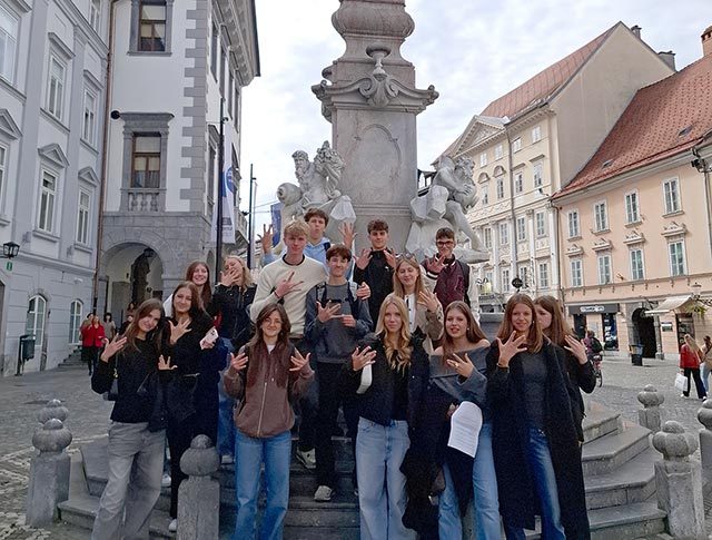 Skupina 15 dijakov stoji na stopnicah Robbovega vodnjaka v starem delu Ljubljane. Iztegnjene prste na rokah držijo v posebnem položaju: sredinec in prstanec se stikata, ostali prsti so narazen.