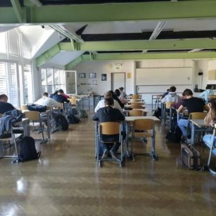 Dijaki sedijo vsak za svojo mizo, ki so postavljene v štiri vrste. Zatopljeni so v reševanje nalog. Vidimo jih v hrbte.