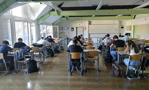 Dijaki sedijo vsak za svojo mizo, ki so postavljene v štiri vrste. Zatopljeni so v reševanje nalog. Vidimo jih v hrbte.