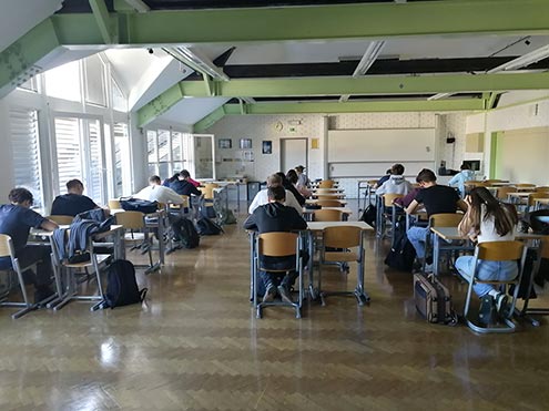 Dijaki sedijo vsak za svojo mizo, ki so postavljene v štiri vrste. Zatopljeni so v reševanje nalog. Vidimo jih v hrbte.