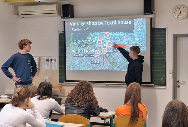 Dva dijaka pred razredom na projekcijskem platnu kažeta, kje v Ljubljani so trgovine z rabljenimi oblačili.