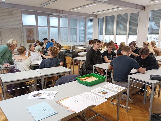 Dijaki sedijo v skupinah po štirje okrog miz v učilnici. Pripravljajo se na skupinsko delo.