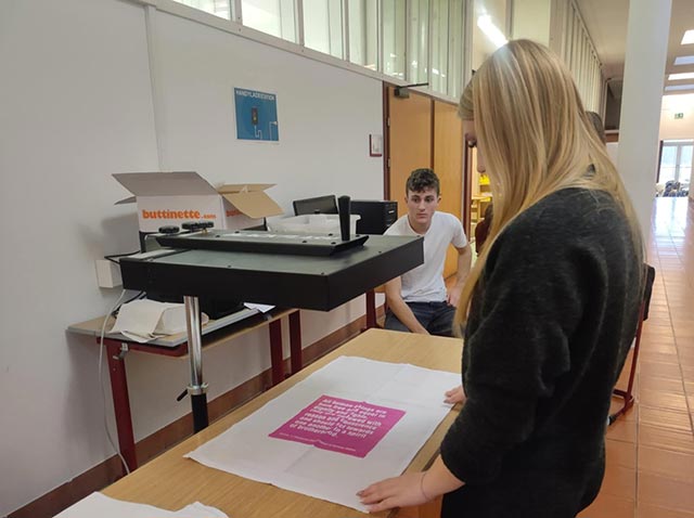 Na mizi je razgrnjen kos blaga z natisnjenim motivom, poleg mize pa sitotiskarska preša. Dijak in dijakinja stojita ob mizi.