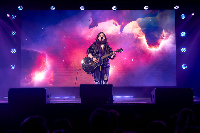 Prizor s prireditve: Na odru stoji dijakinja z akustično kitaro in poje.