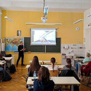 Potopisno predavanje o Afganistanu: Predavatelj živahno govori o fotografiji, ki je prikazana na projekcijskem platnu. Dijaki predavanje spremljajo iz šolskih klopi.