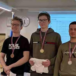 Zmagovalci državnega prvenstva v šahu. Trije fantje stojijo na zmagovalnih stopničha, vsak ima okrog vratu medaljo, ki jo je dosegel. Na obrazih imajo nasmeh.