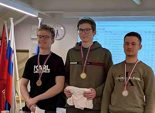 Zmagovalci državnega prvenstva v šahu. Trije fantje stojijo na zmagovalnih stopničha, vsak ima okrog vratu medaljo, ki jo je dosegel. Na obrazih imajo nasmeh.