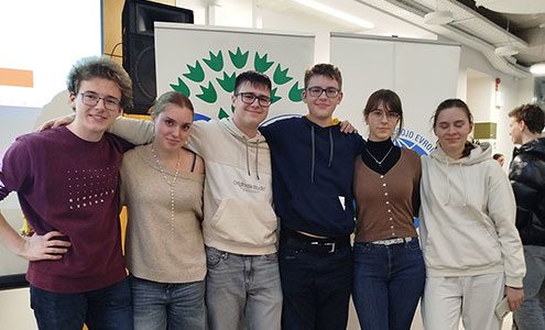 Tri dijakinje in trije dijaki stojijo pred panojem z znakom Ekošole. So nasmejani, objemajo se čez ramena.