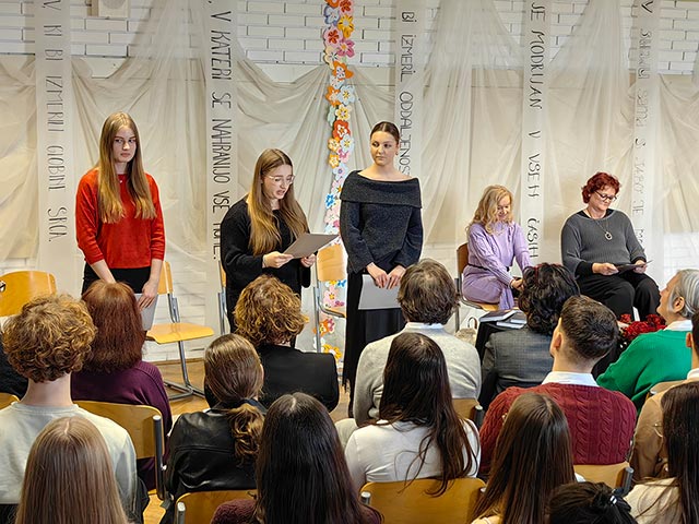 Tri dijakinje stojijo pred občinstvom. Ena od njih interpretira pesem Dragice Čarna. V ozadju fotografije jih opazuje pesnica.