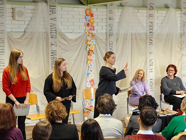 Tri dijakinje stojijo pred občinstvom. Ena od njih interpretira pesem Dragice Čarna. V ozadju fotografije jih opazuje pesnica.