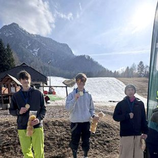 S soncem obsijana gorska pokrajina, v ozadju je s snegom prekrita smučarska proga. V ospredju stojijo trije tekmovalci s kolajnami okrog vratu.