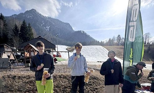 S soncem obsijana gorska pokrajina, v ozadju je s snegom prekrita smučarska proga. V ospredju stojijo trije tekmovalci s kolajnami okrog vratu.