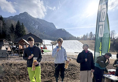 S soncem obsijana gorska pokrajina, v ozadju je s snegom prekrita smučarska proga. V ospredju stojijo trije tekmovalci s kolajnami okrog vratu.