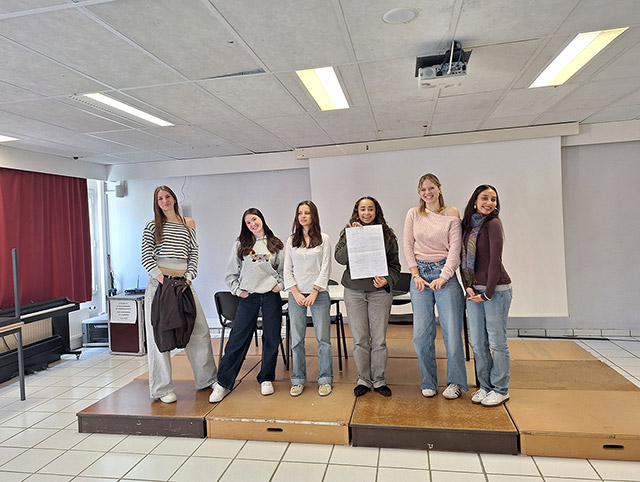 Šest dijakinj stoji druga poleg druge na rahlo privzdignjenem odru v učilnici. Ena od njih v rokah drži velik popisan list paprija. Smejijo se.