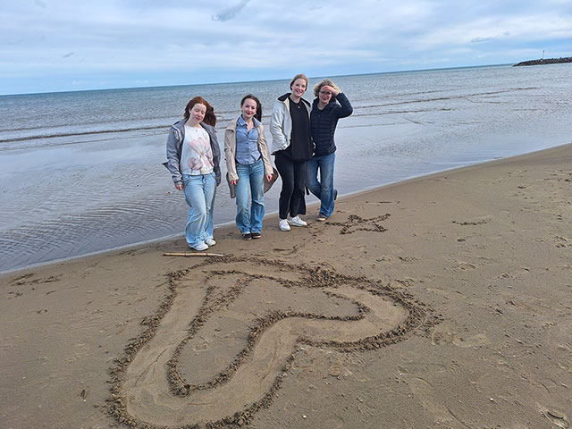 Štiri dijakinje stojijo na peščeni plaži. Je zelo vetrovno, saj jim dolgi lasje in plašči plapolajo v vetru. V pesek so narisale veliko srce in zvezdo. Za njimi je plitvo morje z valovi.