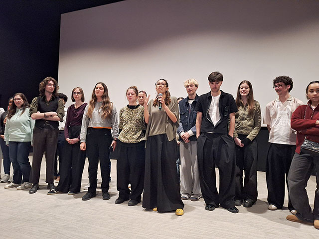 Skupina približno dvajsetih dijakov stoji pred filmskim platnom v kinu. Eno dekle v rokah drži mikrofon in predstavlja film, ki si ga bodo ogledali kasneje.