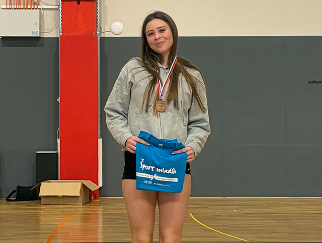 Dijakinja stoji z bronasto medaljo okoli vratu sredi telovadnice. Oblečena je v sivo jopico s kapuco in črne športne kratke hlače. V rokah pred seboj ponosno drži zloženo modro majico z belim napisom "Šport mladih". V ozadju fotografije je siva oblazinjena stena in rdeč steber. Fotografija je bila posneta v telovadnici.
