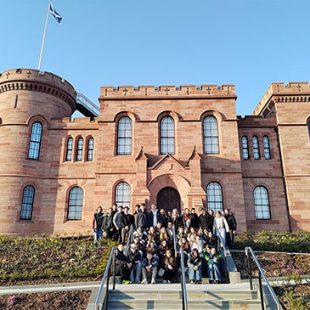 Skupina približno 40 dijakov je razporejena po stopnicah in klančini pred vhodom v grad. Oblečeni so v sproščena oblačila. Stojijo na urejenem območju z nizkim rastlinjem in cvetjem na klančini. Nebo je popolnoma jasno, kar nakazuje na sončen dan. V ozadju je grad Inverness na Škotskem. Zgrajen je iz rdečega kamna. Na levi strani je okrogel stolp, na vrhu katerega na drogu plapola modra škotska zastava z belim križem. Grad ima obokana okna in nazobčan vrh obzidja.