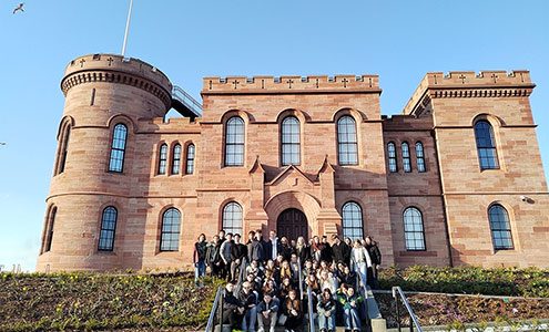 Skupina približno 40 dijakov je razporejena po stopnicah in klančini pred vhodom v grad. Oblečeni so v sproščena oblačila. Stojijo na urejenem območju z nizkim rastlinjem in cvetjem na klančini. Nebo je popolnoma jasno, kar nakazuje na sončen dan. V ozadju je grad Inverness na Škotskem. Zgrajen je iz rdečega kamna. Na levi strani je okrogel stolp, na vrhu katerega na drogu plapola modra škotska zastava z belim križem. Grad ima obokana okna in nazobčan vrh obzidja.