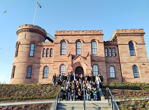 Skupina približno 40 dijakov je razporejena po stopnicah in klančini pred vhodom v grad. Oblečeni so v sproščena oblačila. Stojijo na urejenem območju z nizkim rastlinjem in cvetjem na klančini. Nebo je popolnoma jasno, kar nakazuje na sončen dan. V ozadju je grad Inverness na Škotskem. Zgrajen je iz rdečega kamna. Na levi strani je okrogel stolp, na vrhu katerega na drogu plapola modra škotska zastava z belim križem. Grad ima obokana okna in nazobčan vrh obzidja.