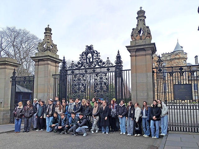 Na sliki je približno 40 dijakov in dijakinj, ki so oblečeni v topla oblačila, kot so bunde, šali in kavbojke. Nekaj jih stoji, nekaj pa jih čepi v ospredju. V ozadju so mogočna, črna kovana vrata z bogatim okrasjem. Vrata so vpeta med masivne kamnite stebre, na vrhu katerih so skulpture s kronami. Desno za vrati se vidi del kamnite zgradbe s stolpičem in koničasto streho. Gre za palačo Holyroodhouse v Edinburgu. Nebo je oblačno. 