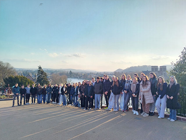 Skupina približno 40 dijakov in dijakinj stoji v dolgi, rahlo ukrivljeni vrsti na široki, asfaltirani razgledni ploščadi. Sonca je nizko na levi strani. Večina je oblečena v športno-elegantna zimska in prehodna oblačila (bunde, puloverji, kavbojke). Za skupino se razprostira panoramski razgled na mesto. Vidi se reka, ki teče skozi dolino, obdana z drevjem in mestnimi stavbami. Na desni strani v daljavi izstopa velika cerkev z dvema stolpoma. V ozadju so vidni mehki griči pod svetlim, rahlo oblačnim nebom.