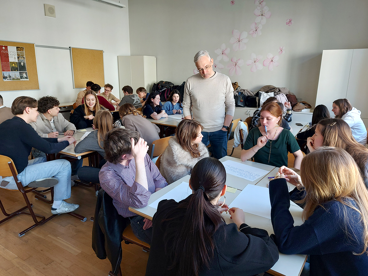 Dijaki v učilnici sedijo v skupinah za mizami in aktivno sodelujejo pri delu. Žiga Valetič stoji ob eni izmed skupin in se z njimi pogovarja. 