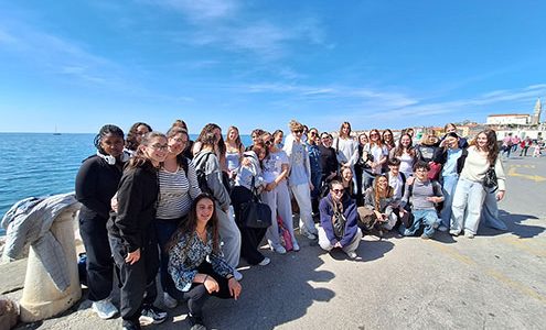 Velika skupina nasmejanih dijakov pozira na obalni ploščadi v Piranu. Nebo je popolnoma jasno. V ozadju se v daljavi vidi veduta Pirana s cerkvenim zvonikom.